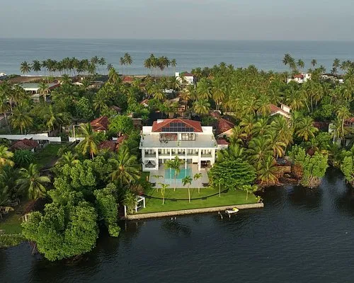 Aerial view of a tropical villa with a swimming pool on a waterway, surrounded by palm trees and greenery.